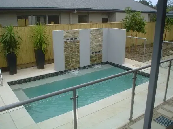 Backyard swimming pool with glass railing, stone water wall, and potted plants