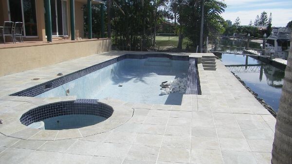 Empty tiled pool deck with a small round spa, beside a canal and palm trees