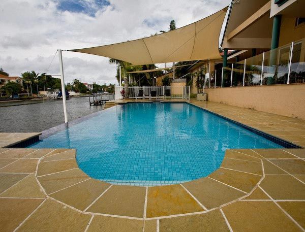 Outdoor swimming pool with blue water, tan stone deck, and beige shade sails beside a modern building