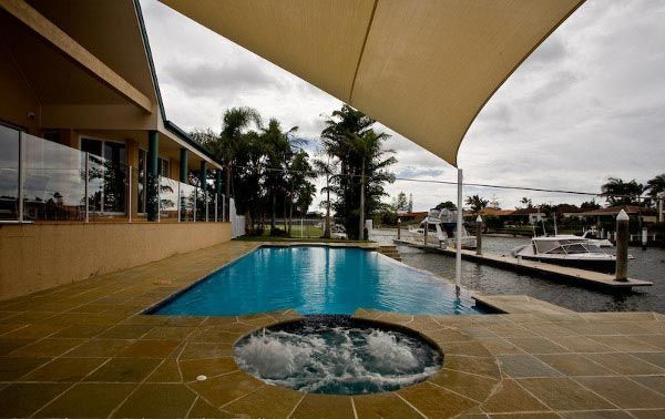 Modern poolside patio with turquoise pool, hot tub, and sail shade beside a waterfront.