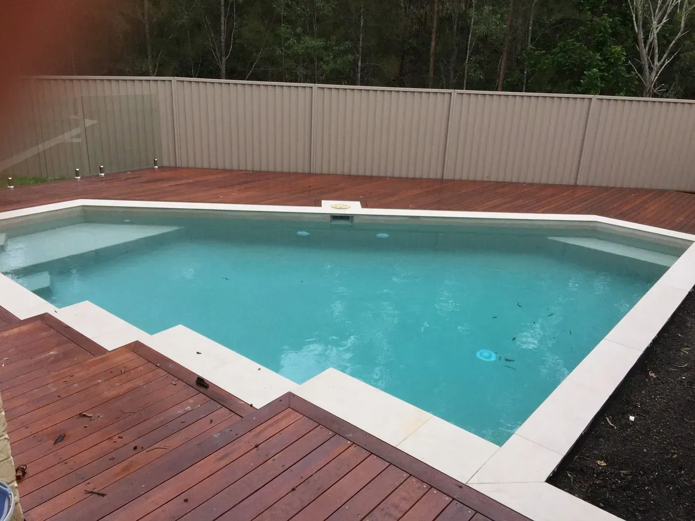 Rectangular outdoor swimming pool with turquoise water, white edging, and wooden deck beside a fence and trees