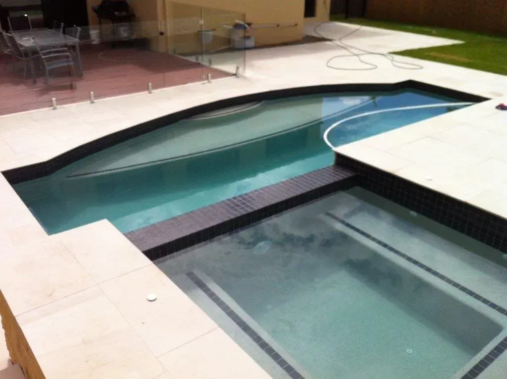 Two-tier backyard pool with turquoise water, white tile deck, and black coping at night