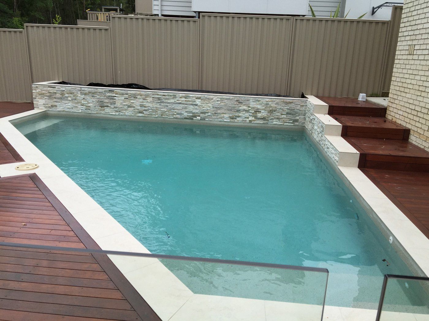 Backyard swimming pool with turquoise water, wooden deck, stone waterfall wall, and glass safety barrier