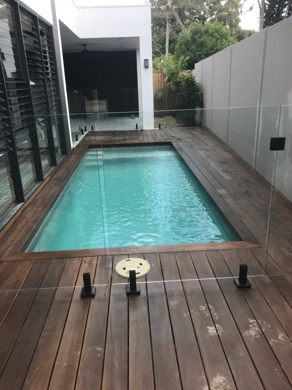 Narrow backyard pool with turquoise water, bordered by wooden decking and a modern house wall.