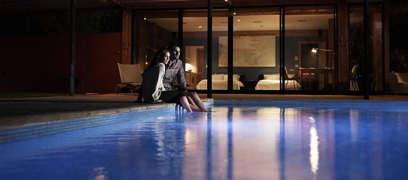 Person sitting by a lit swimming pool at night, with reflections on the blue water and a house in the background