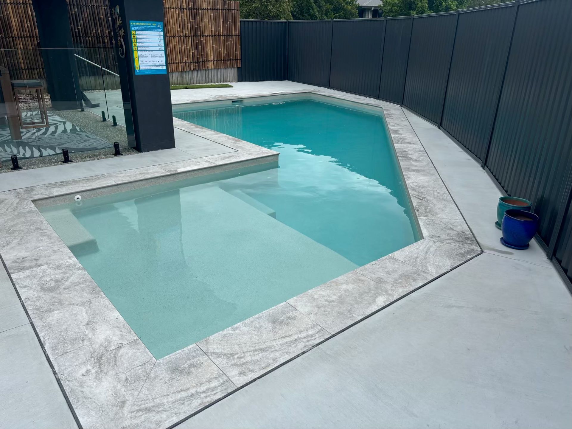 Small backyard swimming pool with pale blue water, white stone deck, and dark fence enclosure