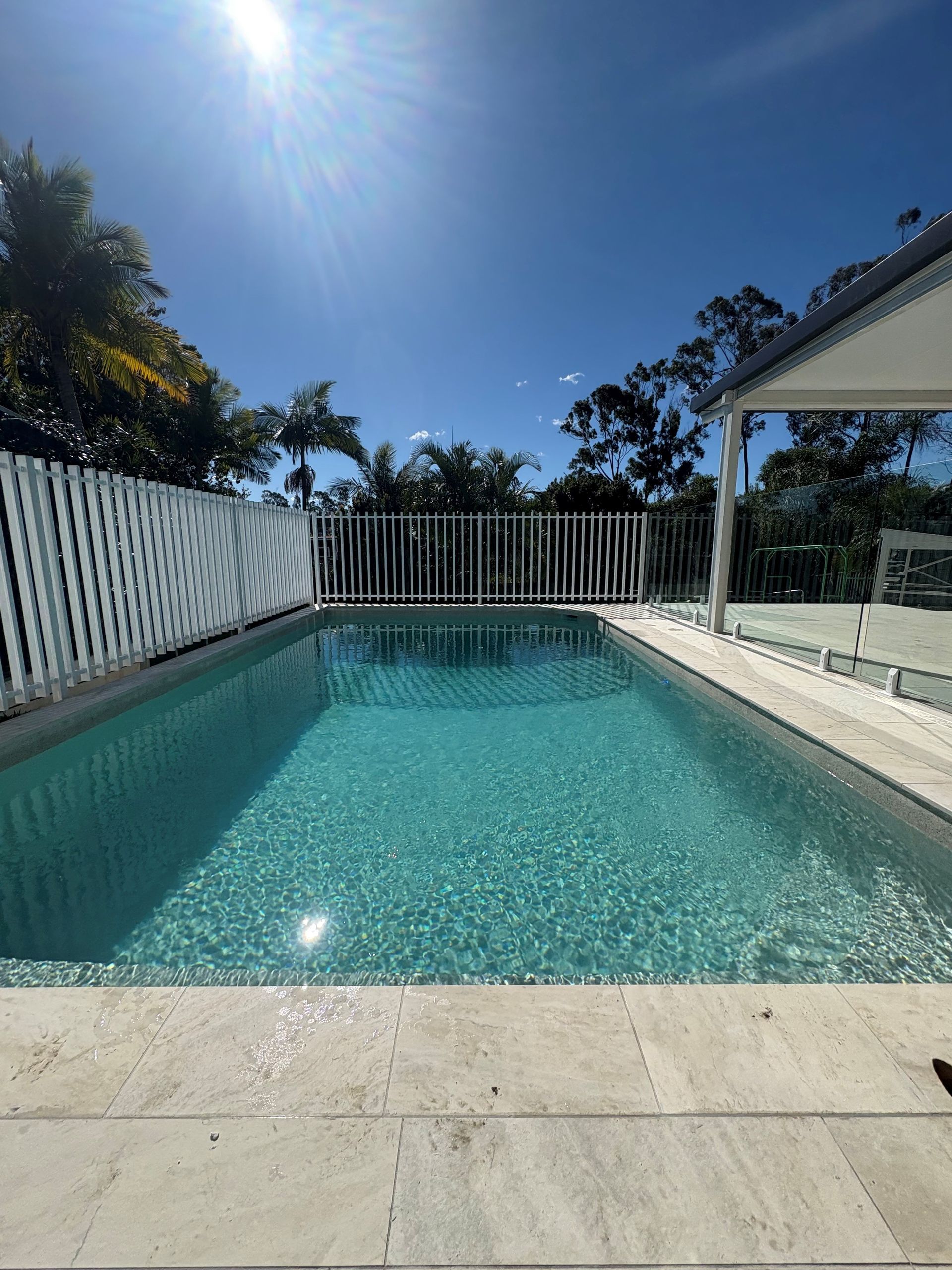Sunny backyard swimming pool with turquoise water, white fence, and patio under a clear blue sky