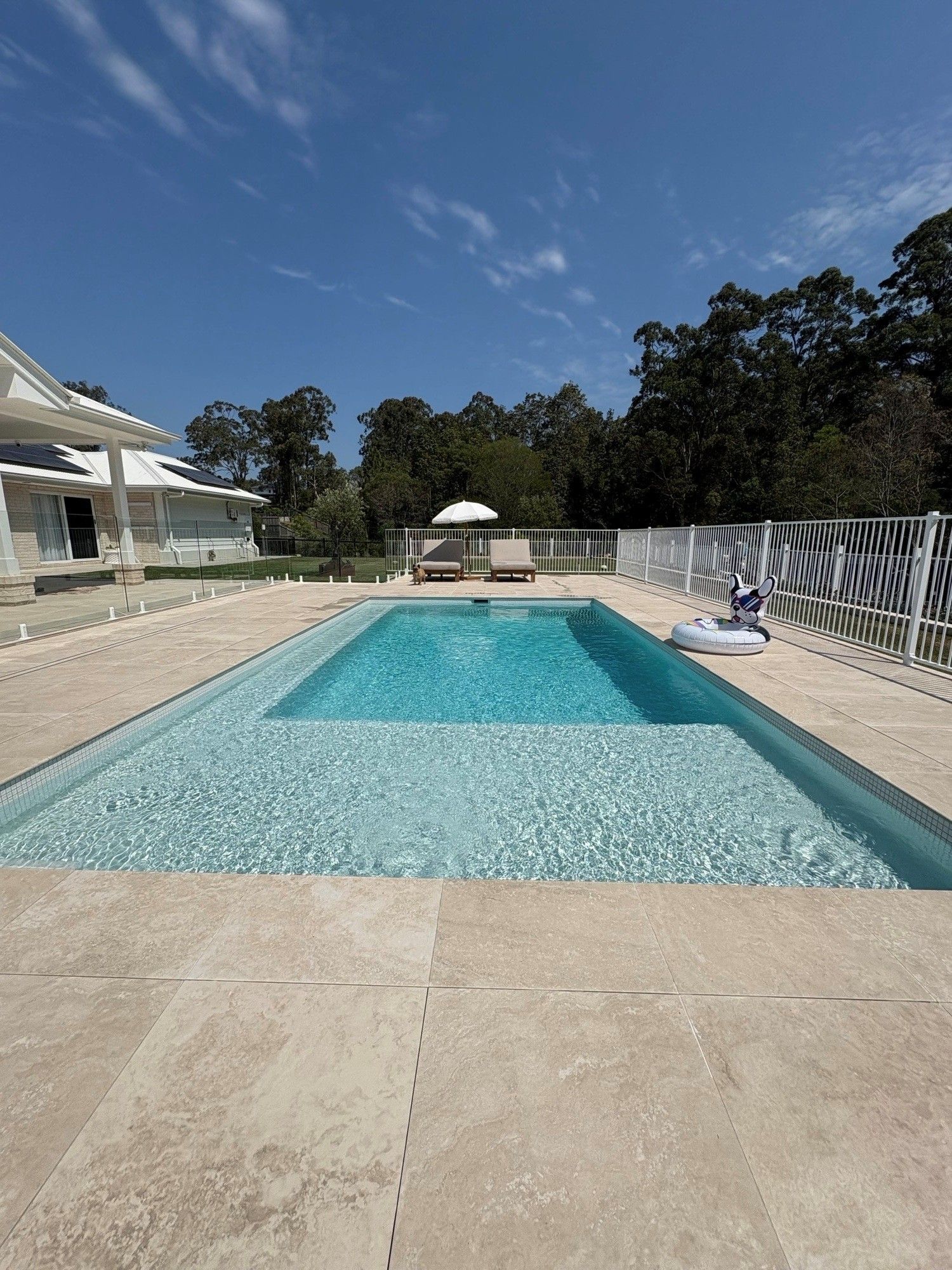 Outdoor swimming pool with turquoise water, stone patio, lounge chairs, and a clear blue sky.
