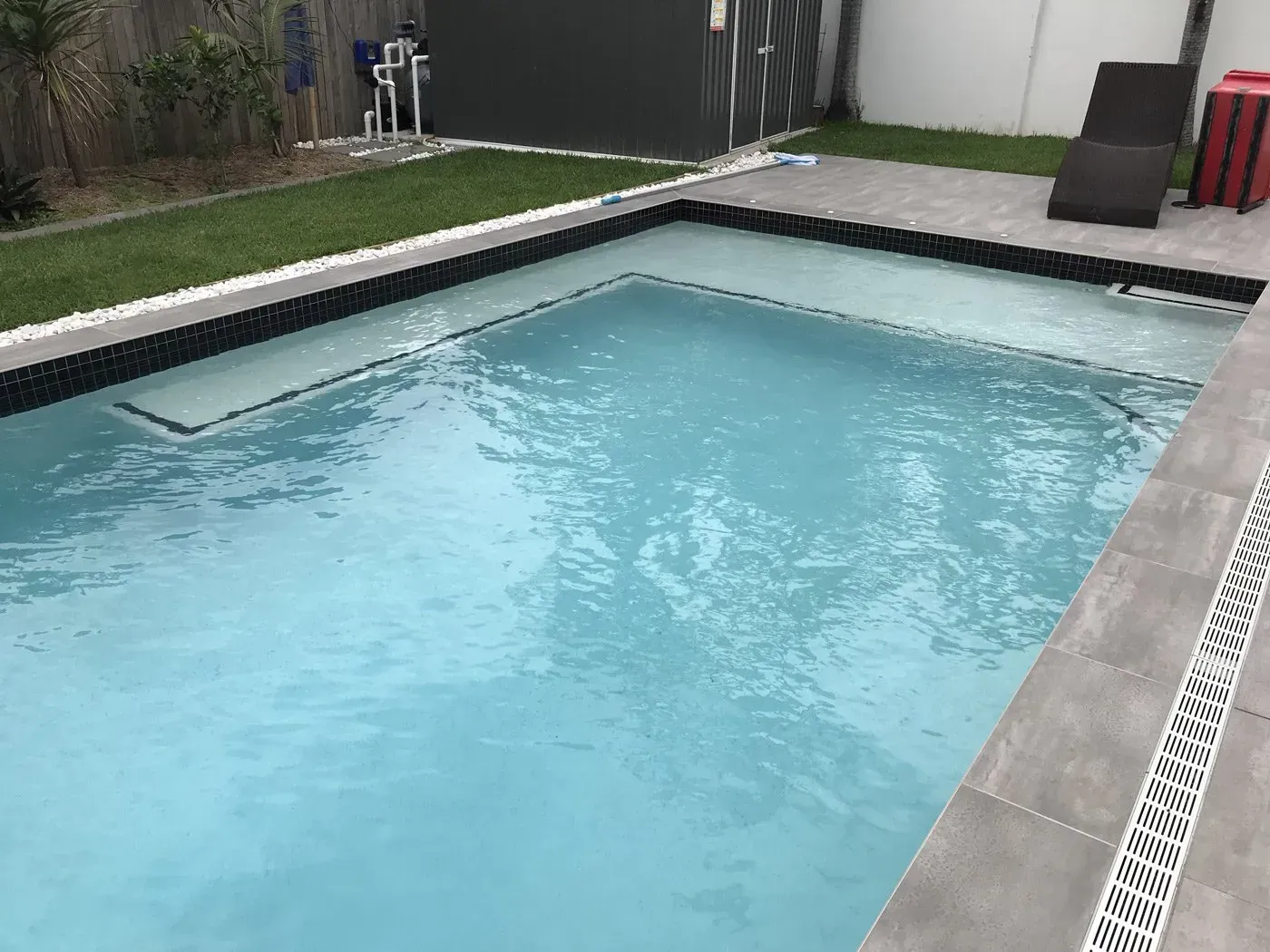 Backyard swimming pool with clear blue water, gray tile deck, and surrounding fence and lawn