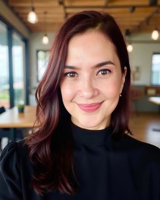 Smiling person with dark hair in a black top, seated in a bright café with blurred lights in the background