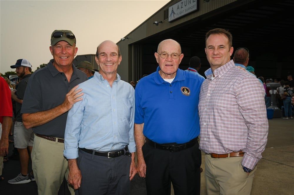 Four men are posing for a picture in front of an airplane hangar.