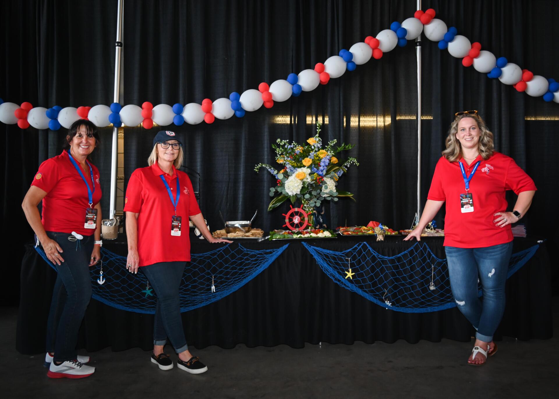 Three women are standing in front of a table with balloons hanging from the ceiling.