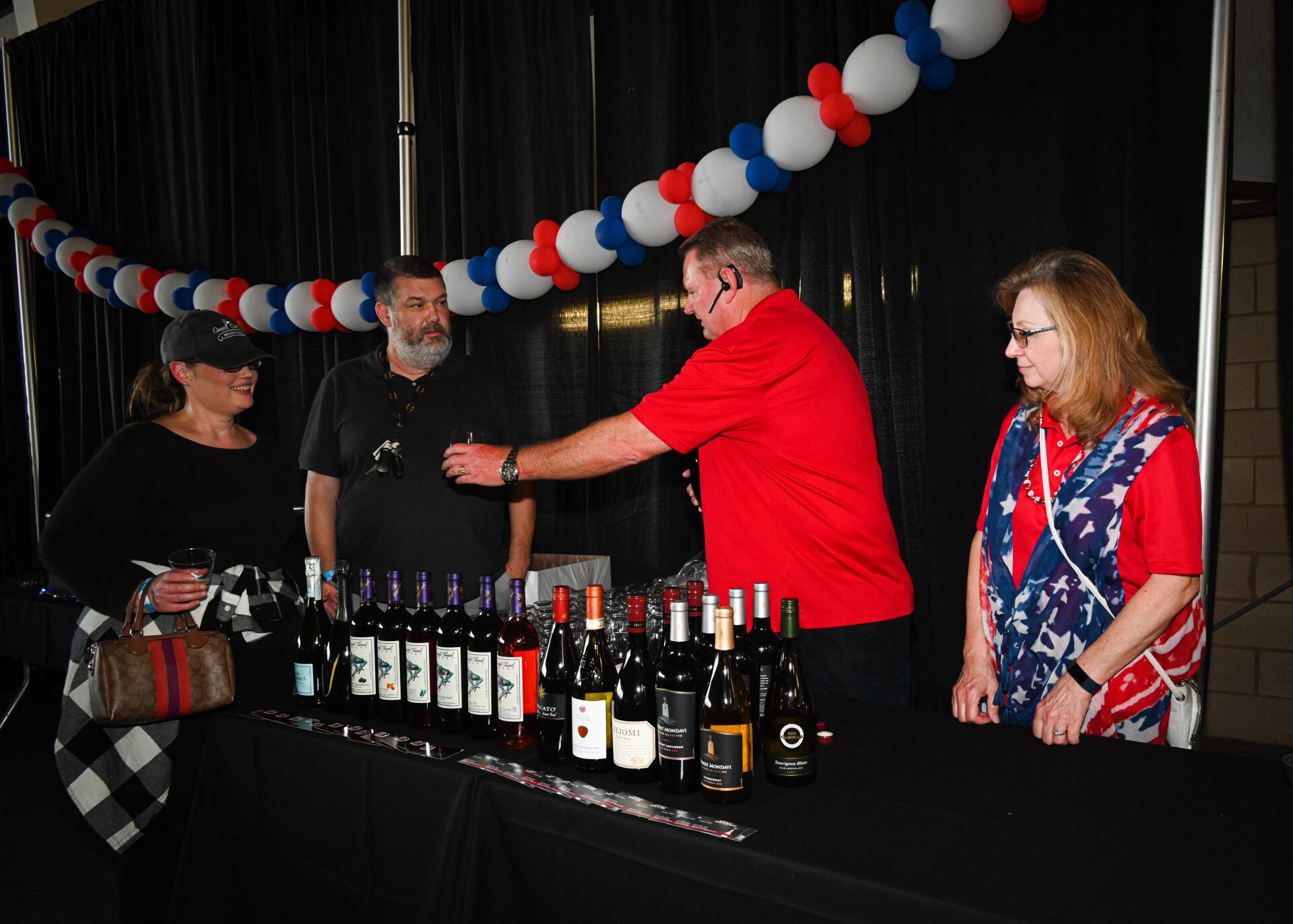 A group of people are standing around a table with bottles of wine on it.