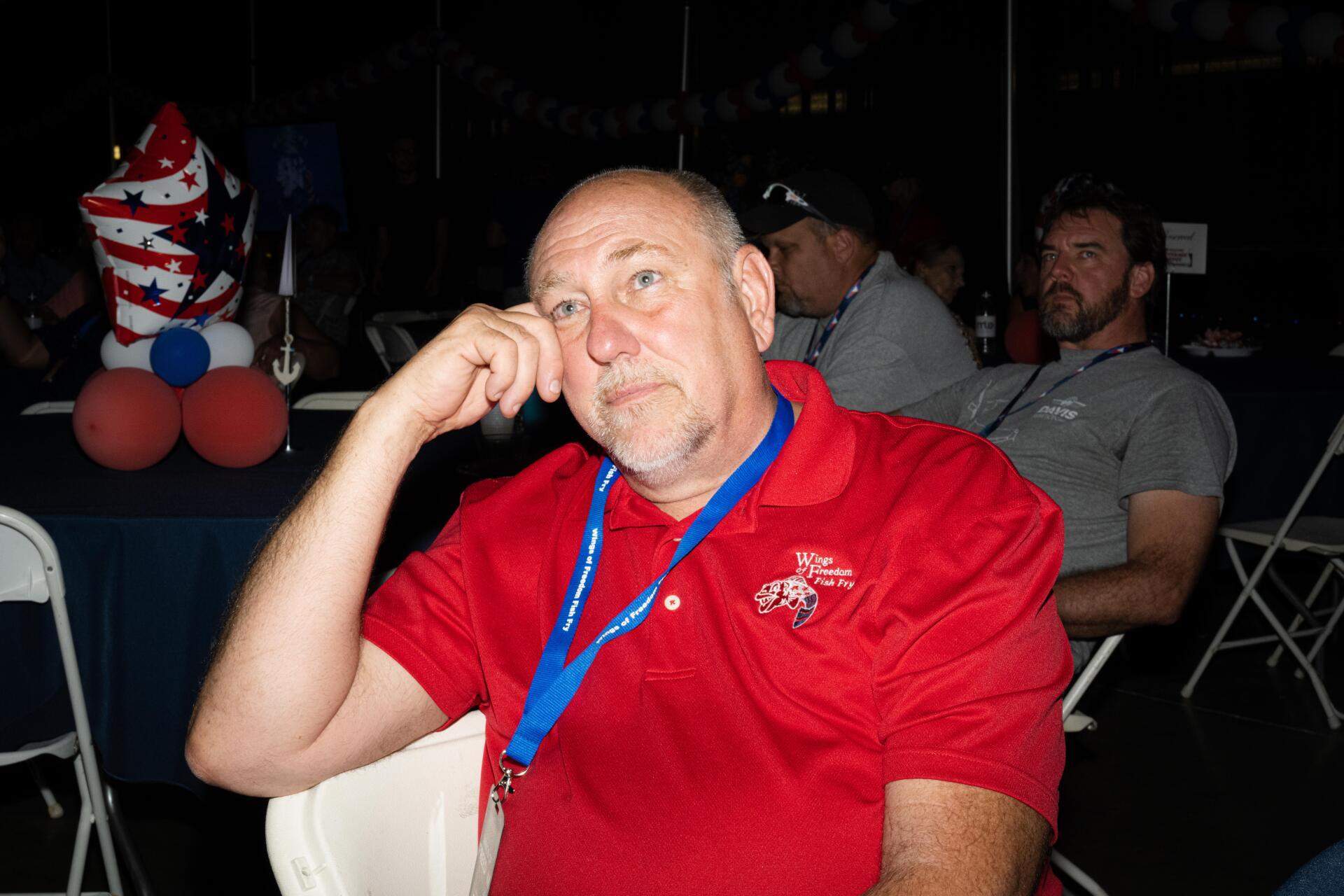 A man in a red shirt is sitting in a chair with his hand on his chin.