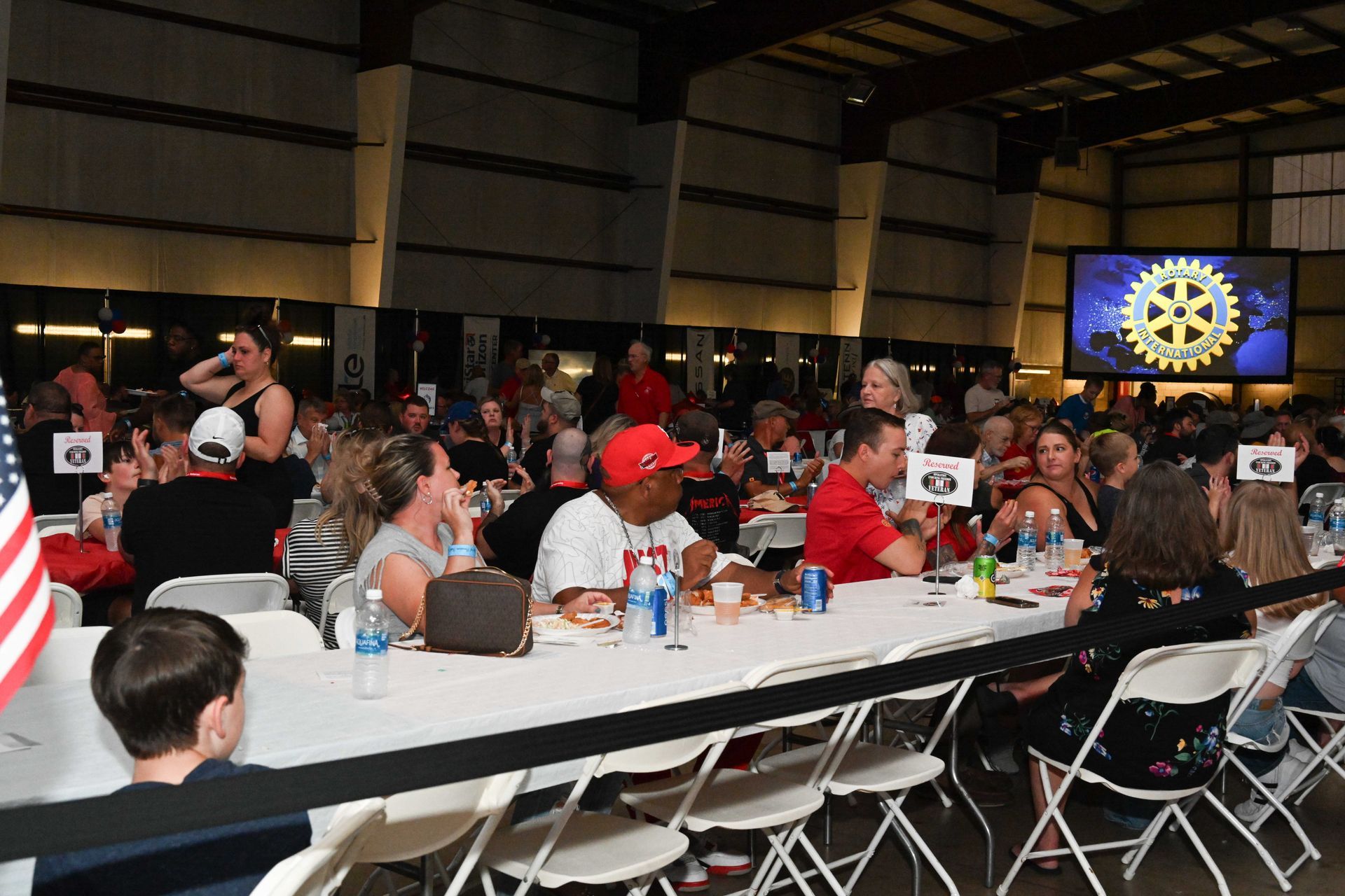 A large group of people are sitting at long tables in front of a large screen with a rotary logo on it.
