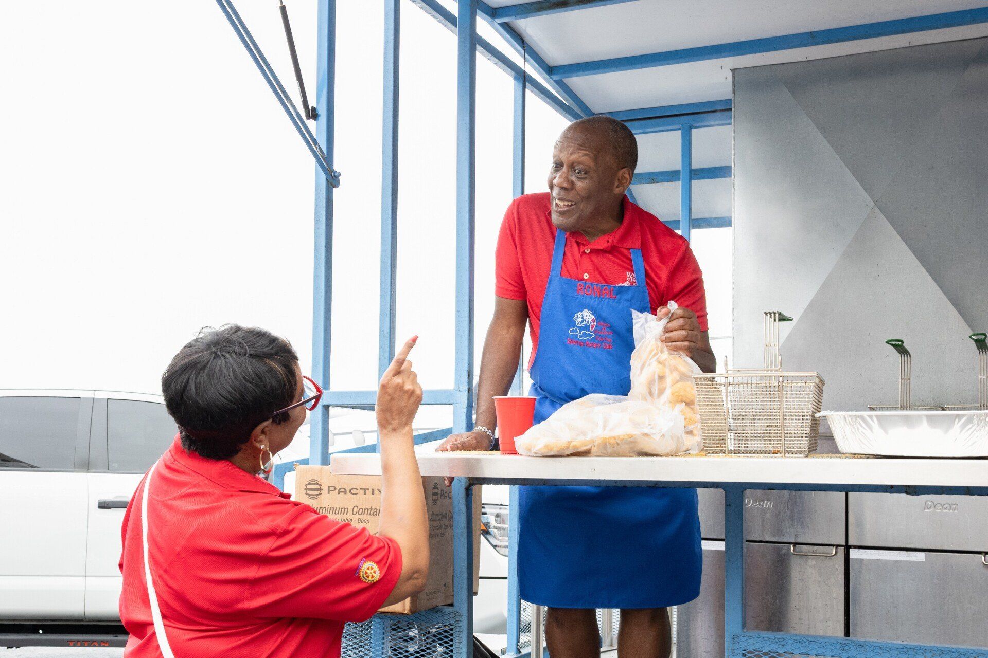 A man in a blue apron is talking to a woman in a red shirt.