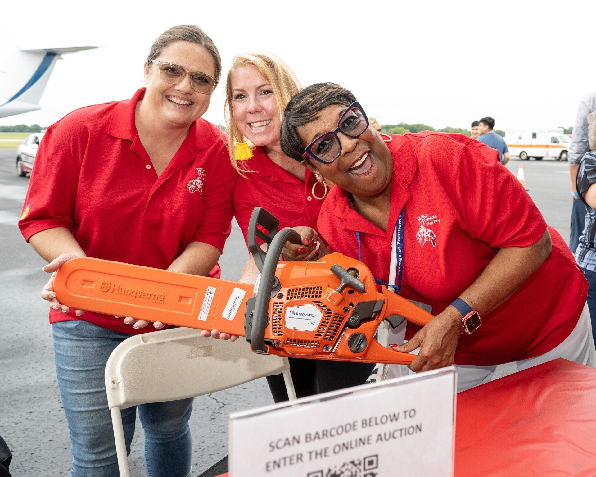 Three women in red shirts are holding a chainsaw and posing for a picture.