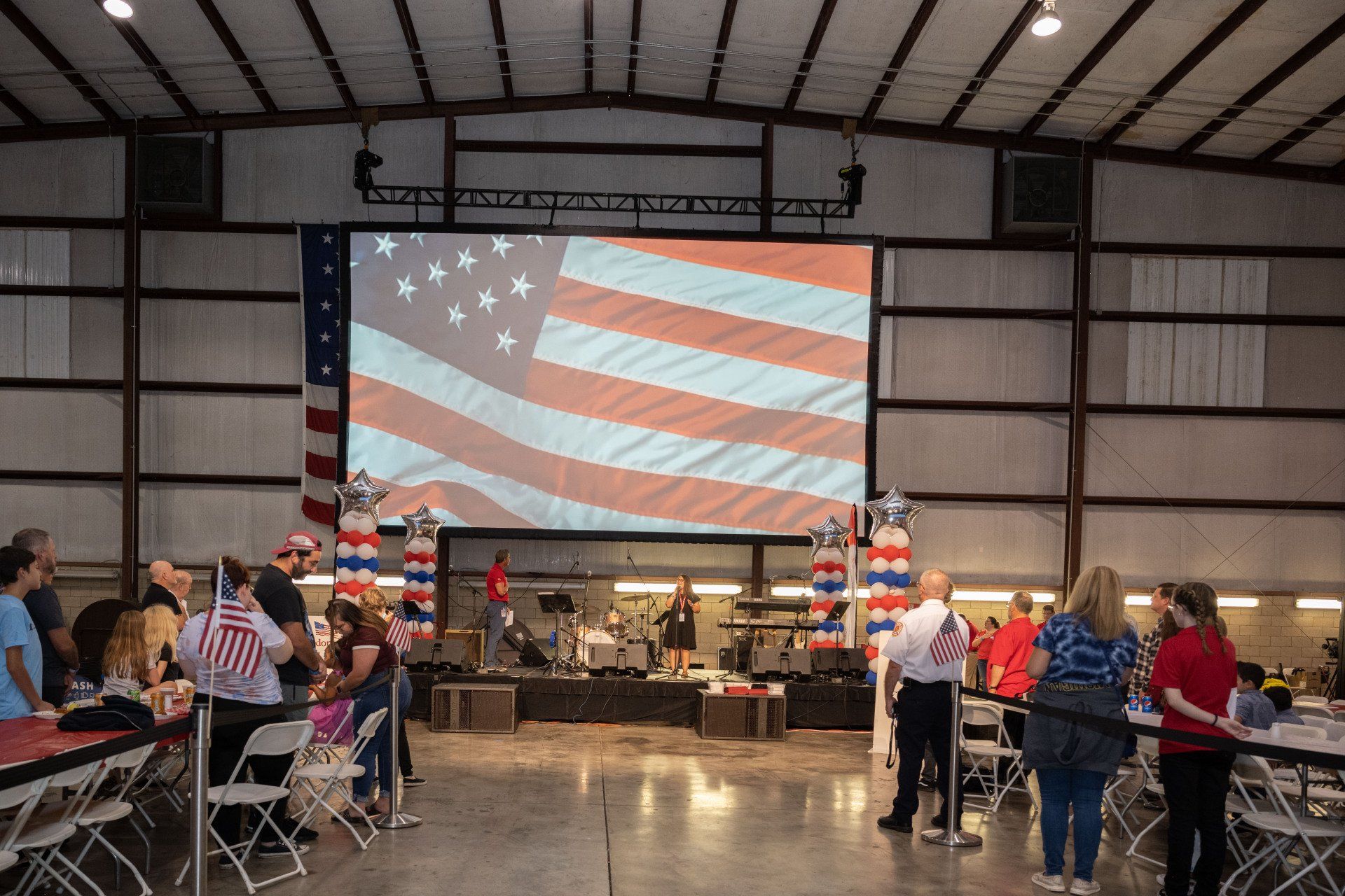 A group of people are sitting at tables in front of a large screen with an american flag on it.