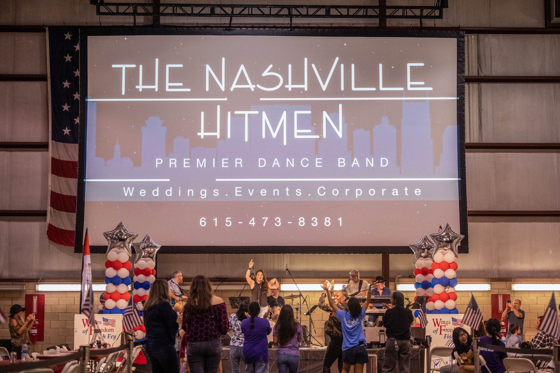 A group of people are standing in front of a large screen that says the nashville hitmen premier dance band