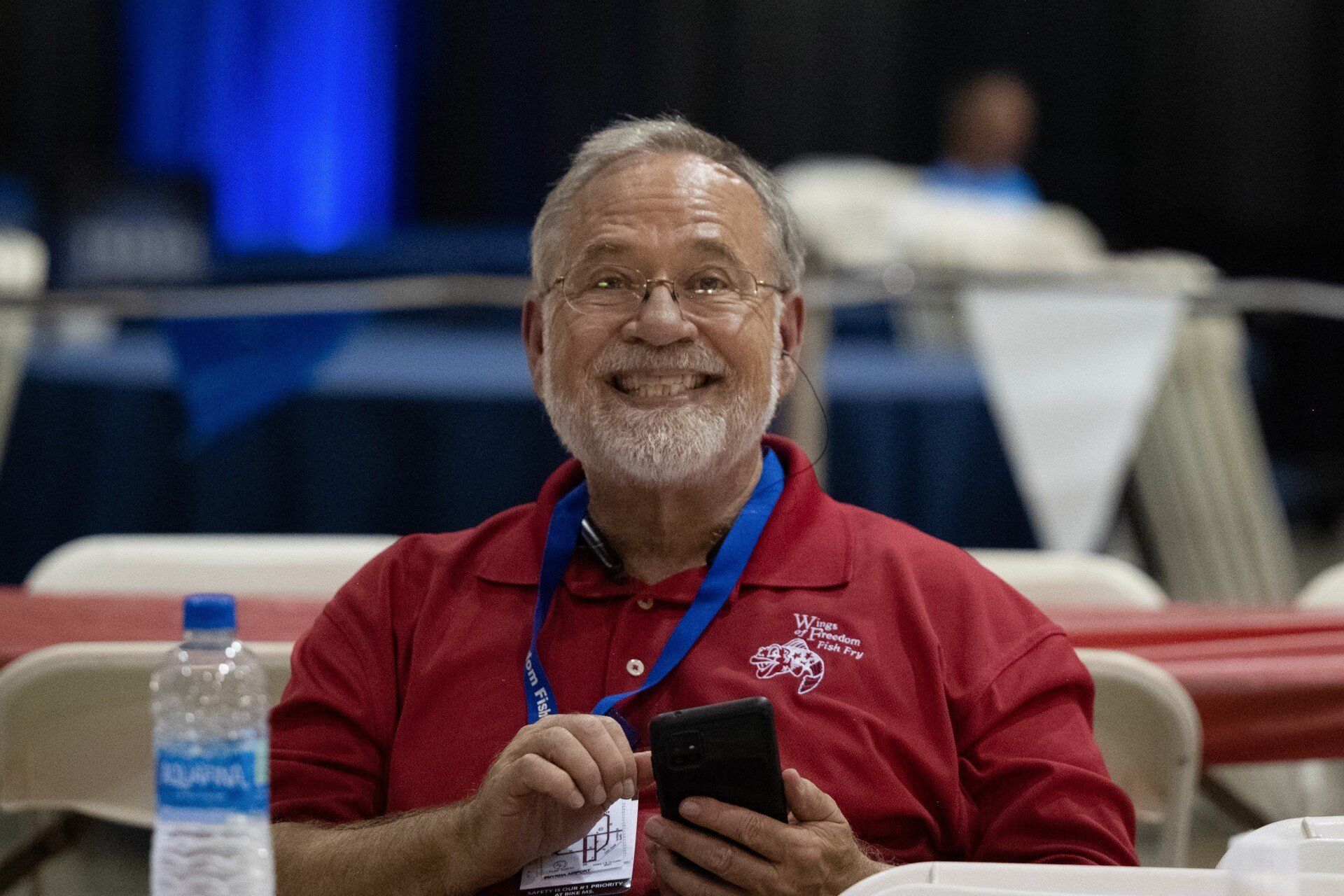 A man in a red shirt is sitting at a table holding a cell phone.