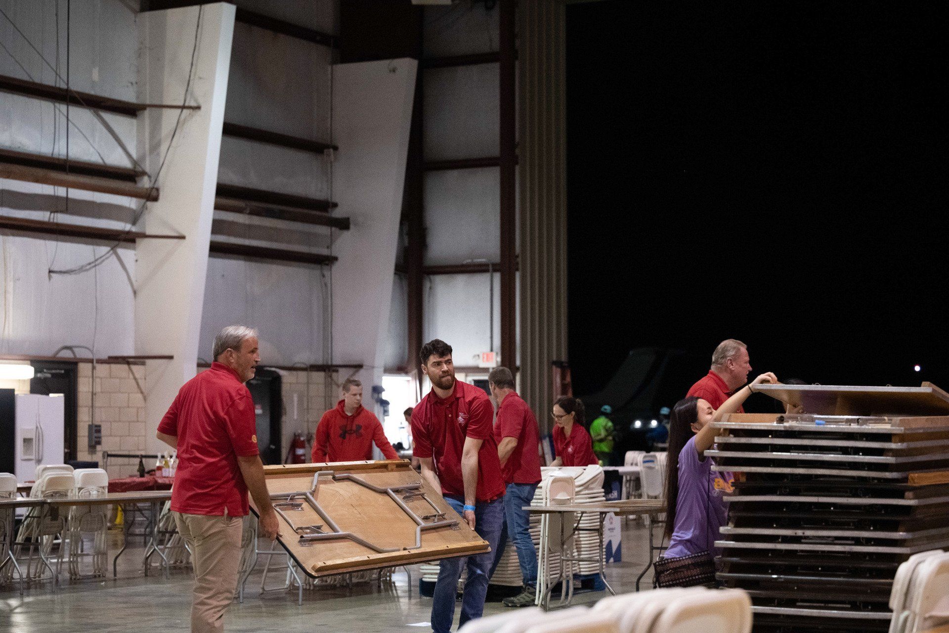 A group of men in red shirts are carrying tables in a warehouse
