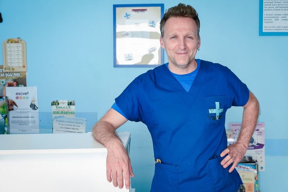 Un uomo in camice blu da medico è in piedi nella sala d'attesa di una clinica, appoggiato a un bancone e sorridente.