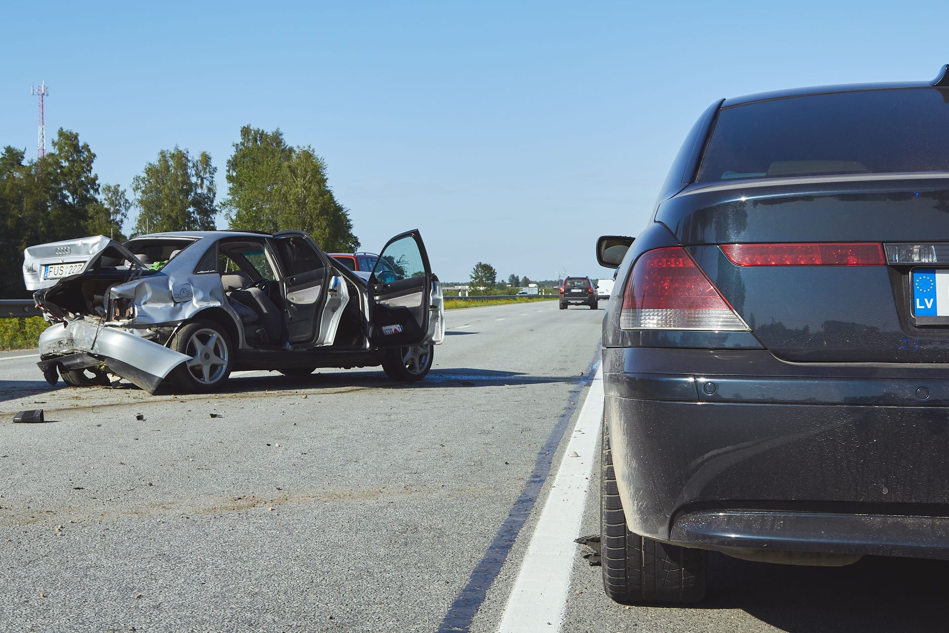 Severe car crash on a highway shoulder, with a damaged black sedan and an open door beside another car.