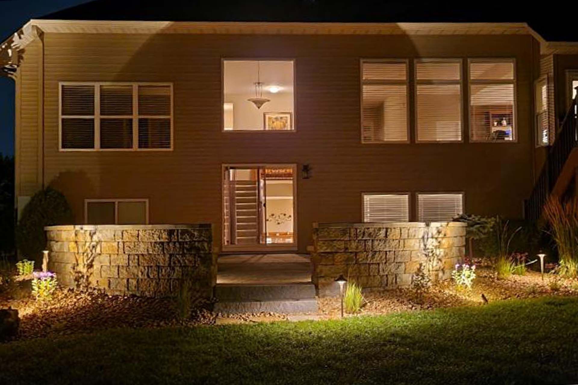 A house with a lot of windows is lit up at night showcasing the retaining wall and patio