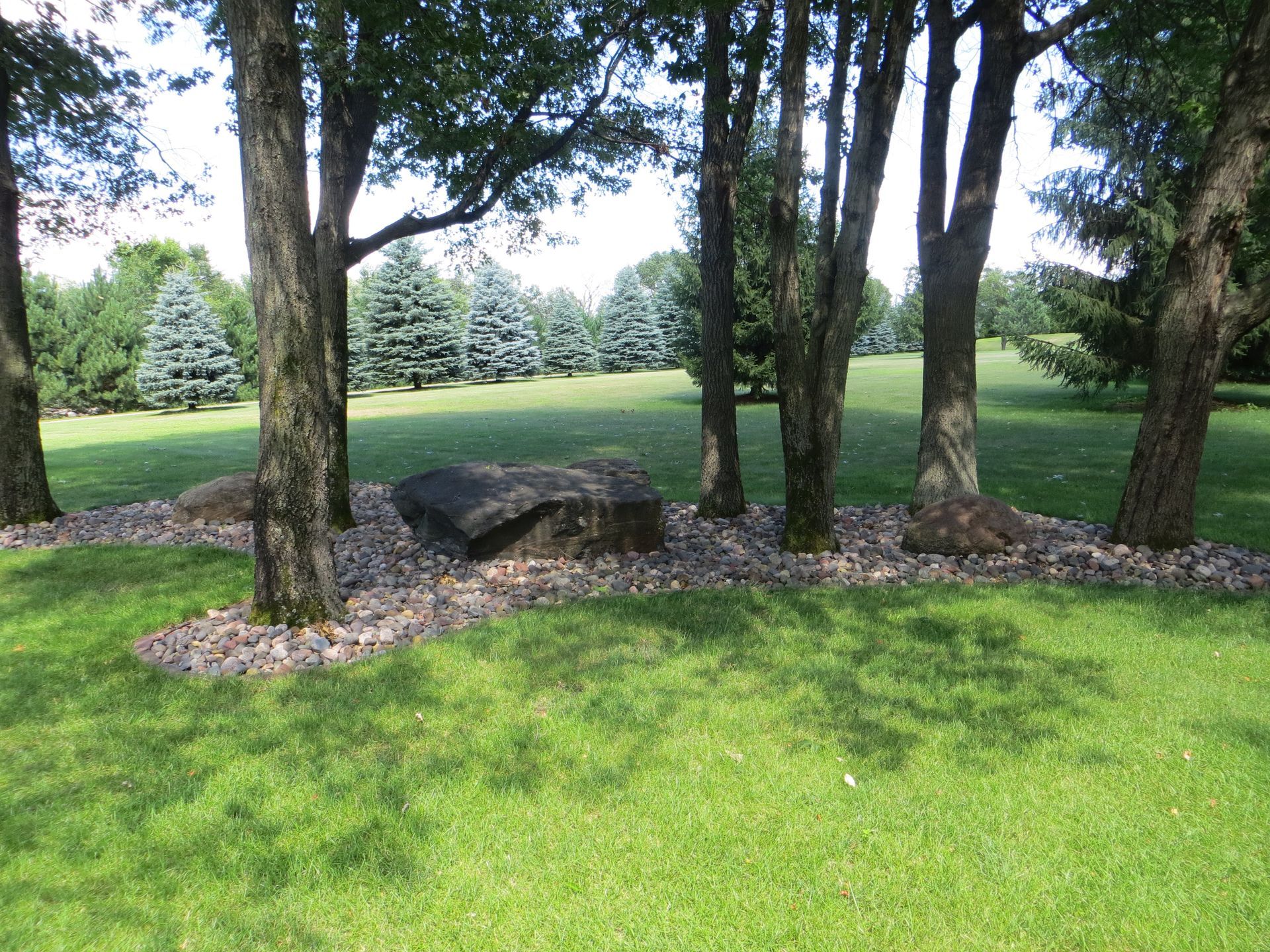 A large rock sits in the middle of a mulch garden surrounded by trees and lush green grass.