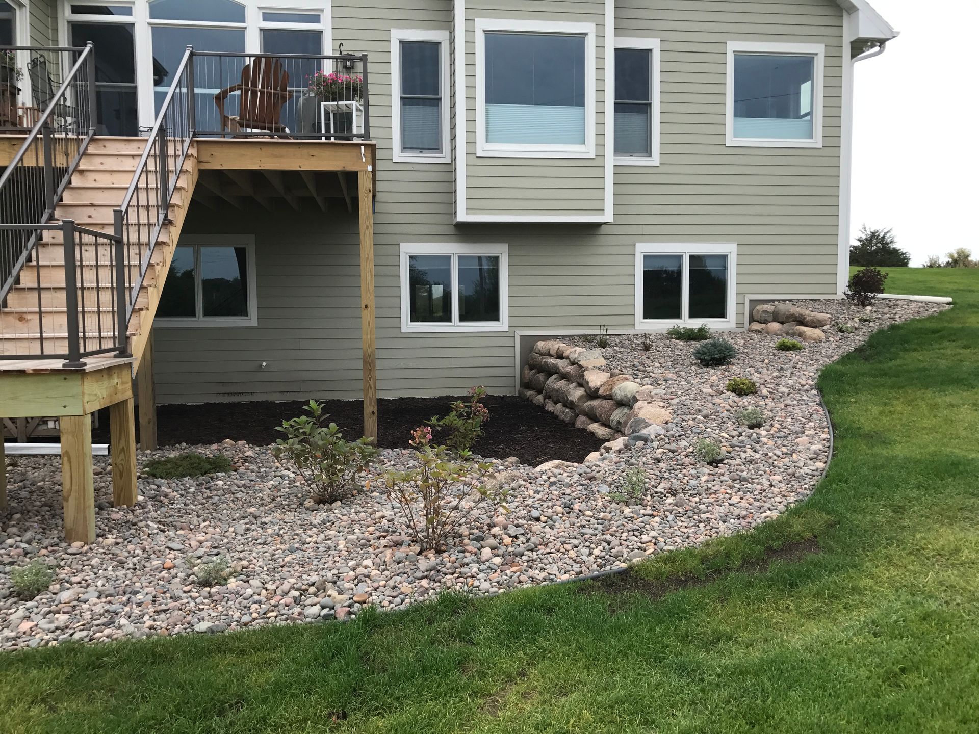 A rock garden with a retaining wall up against the back of a house with a deck and stairs.