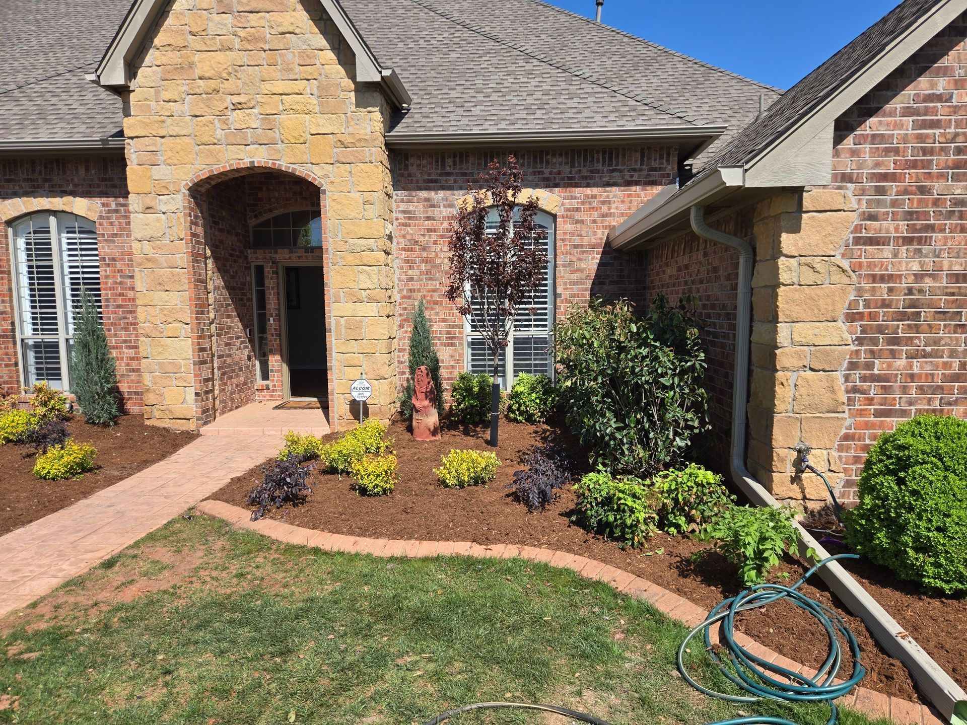 Brick house front yard with landscaped flower beds, walkway, and a garden hose near the entrance