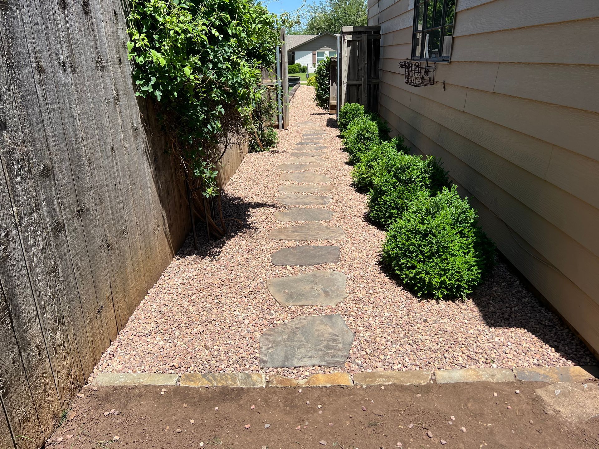 Narrow gravel walkway between a stucco wall and house, with stepping stones and green shrubs.