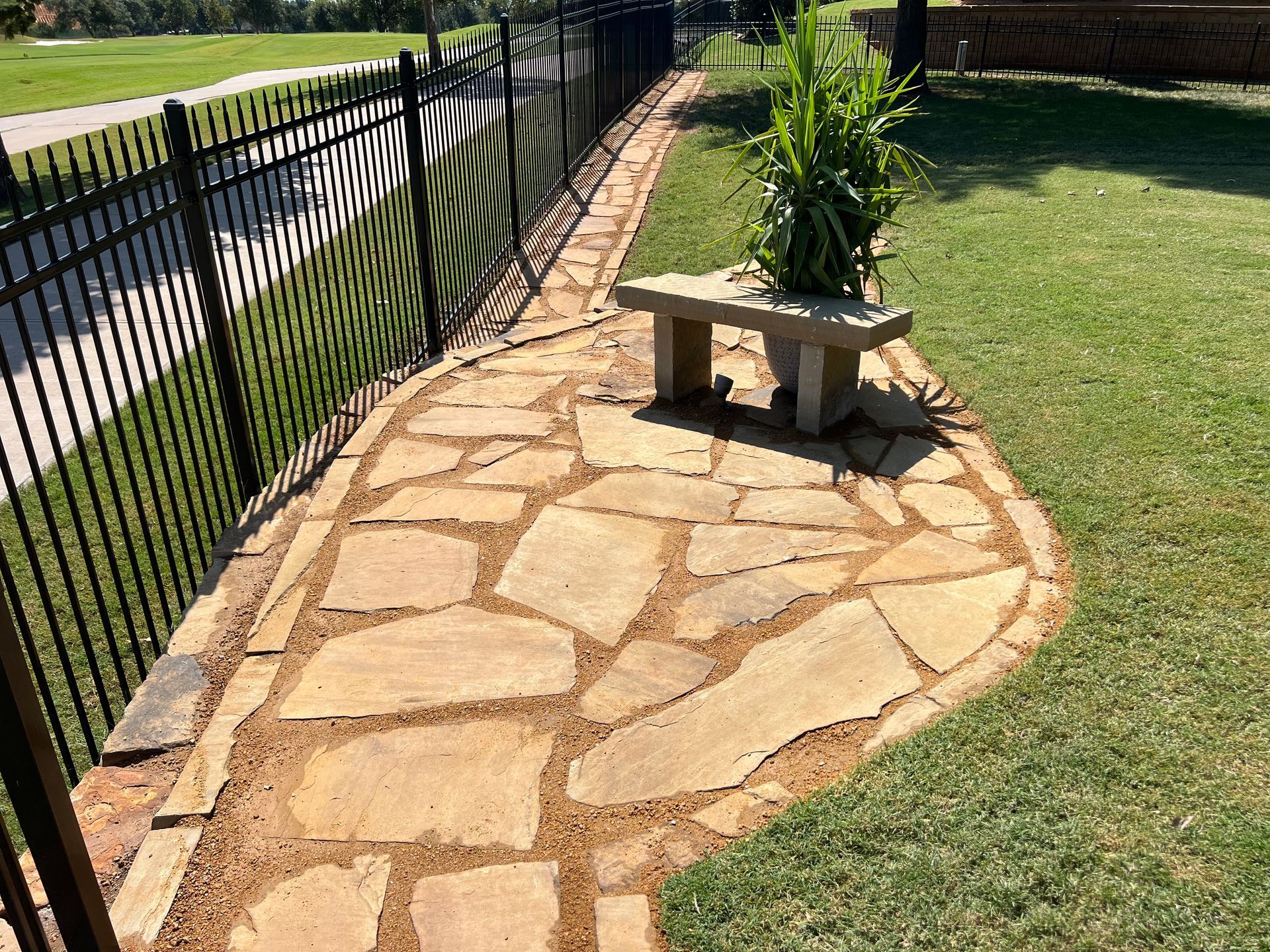 Stone garden path beside black metal fence and grassy lawn, with a stone bench and potted plant.