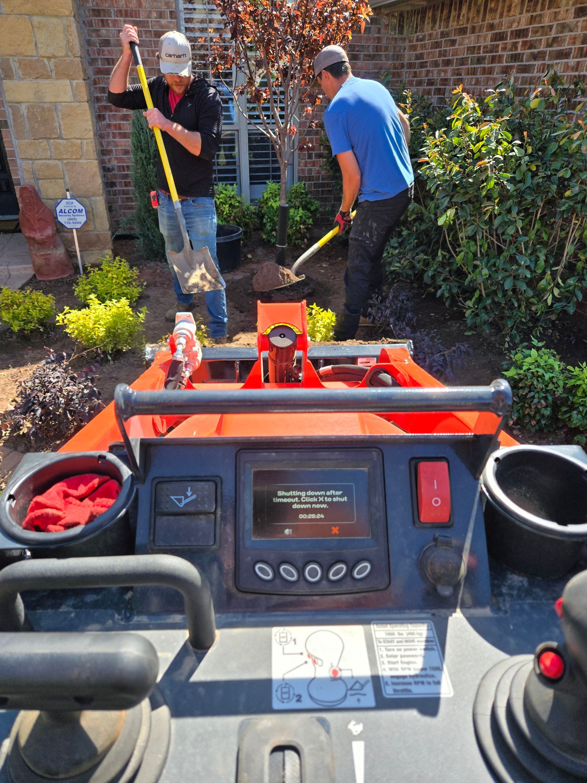 Riding lawn mower view toward two workers landscaping a garden path and brick house