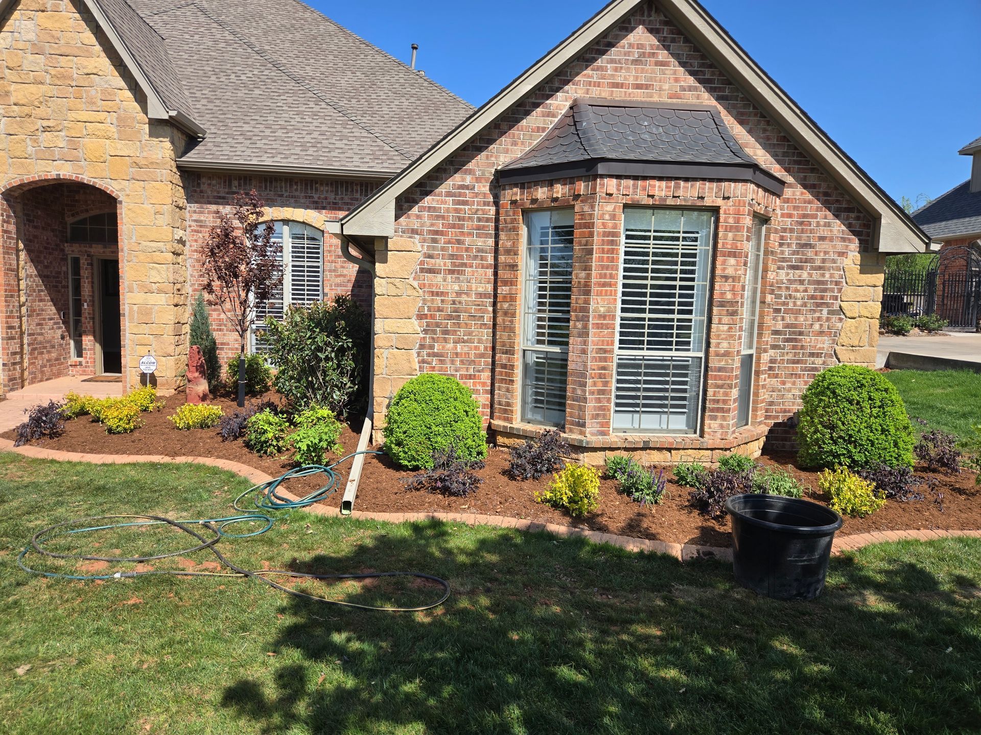 Brick suburban house with front flower beds, trimmed shrubs, and a large windowed gable.