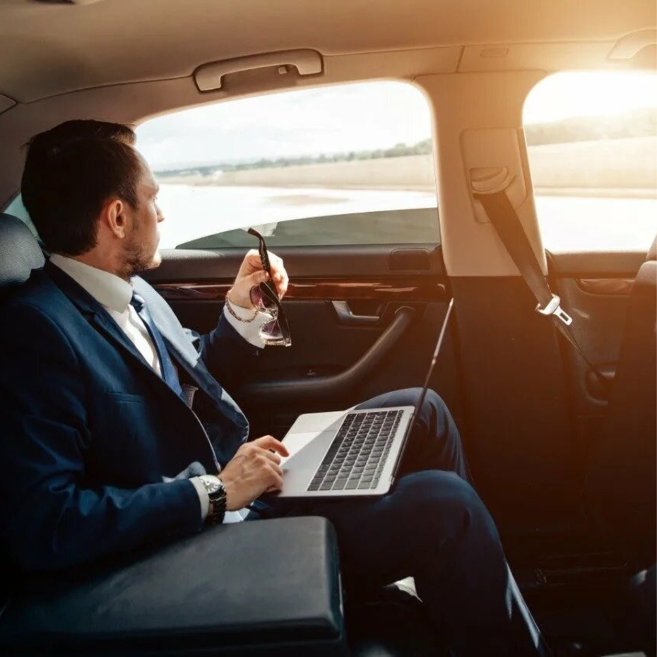 Businessman in a car, using a laptop and holding a phone by the window at sunset