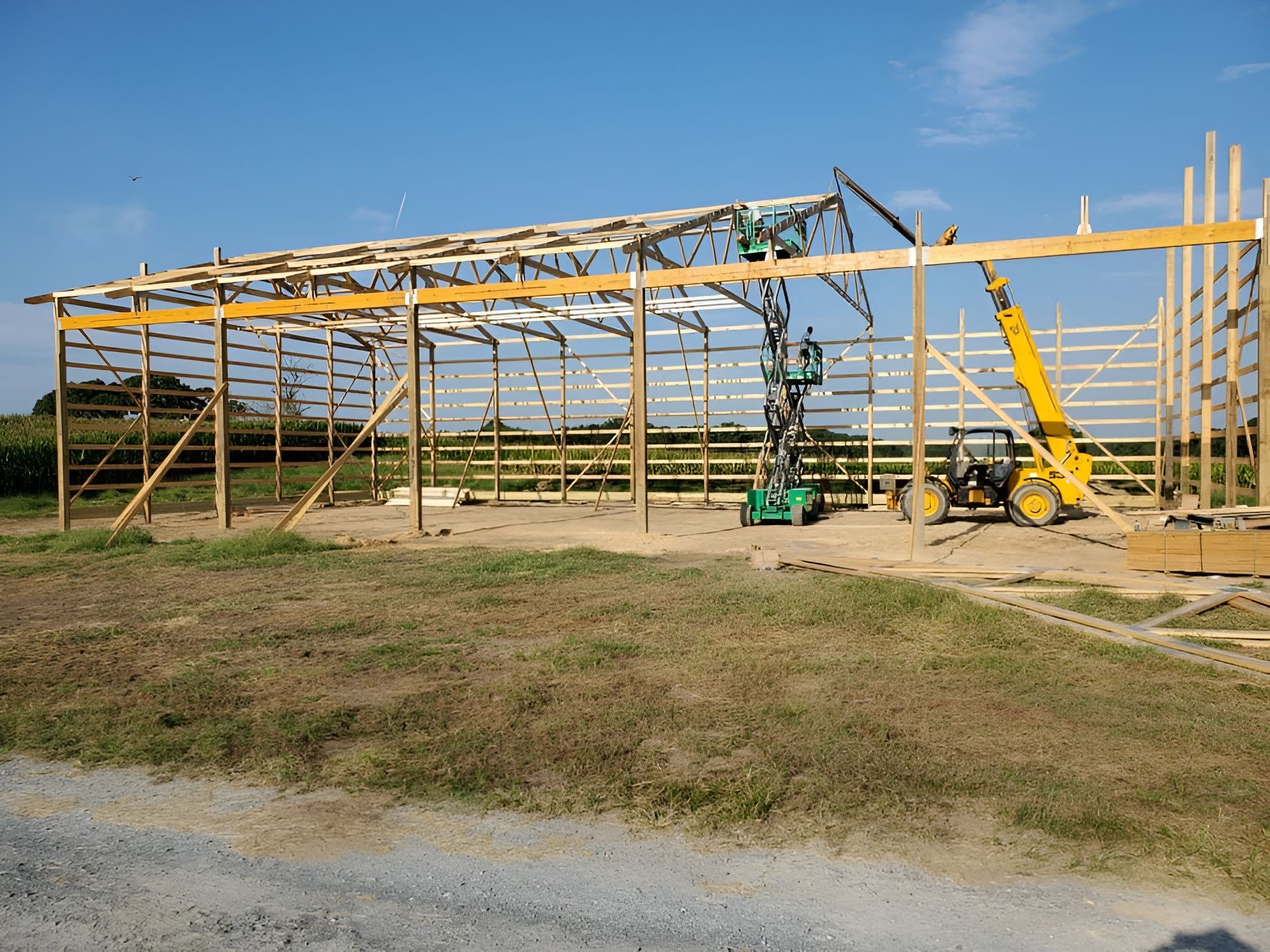 Construction workers use a yellow telehandler and a green scissor lift to frame a large wooden post-frame building.