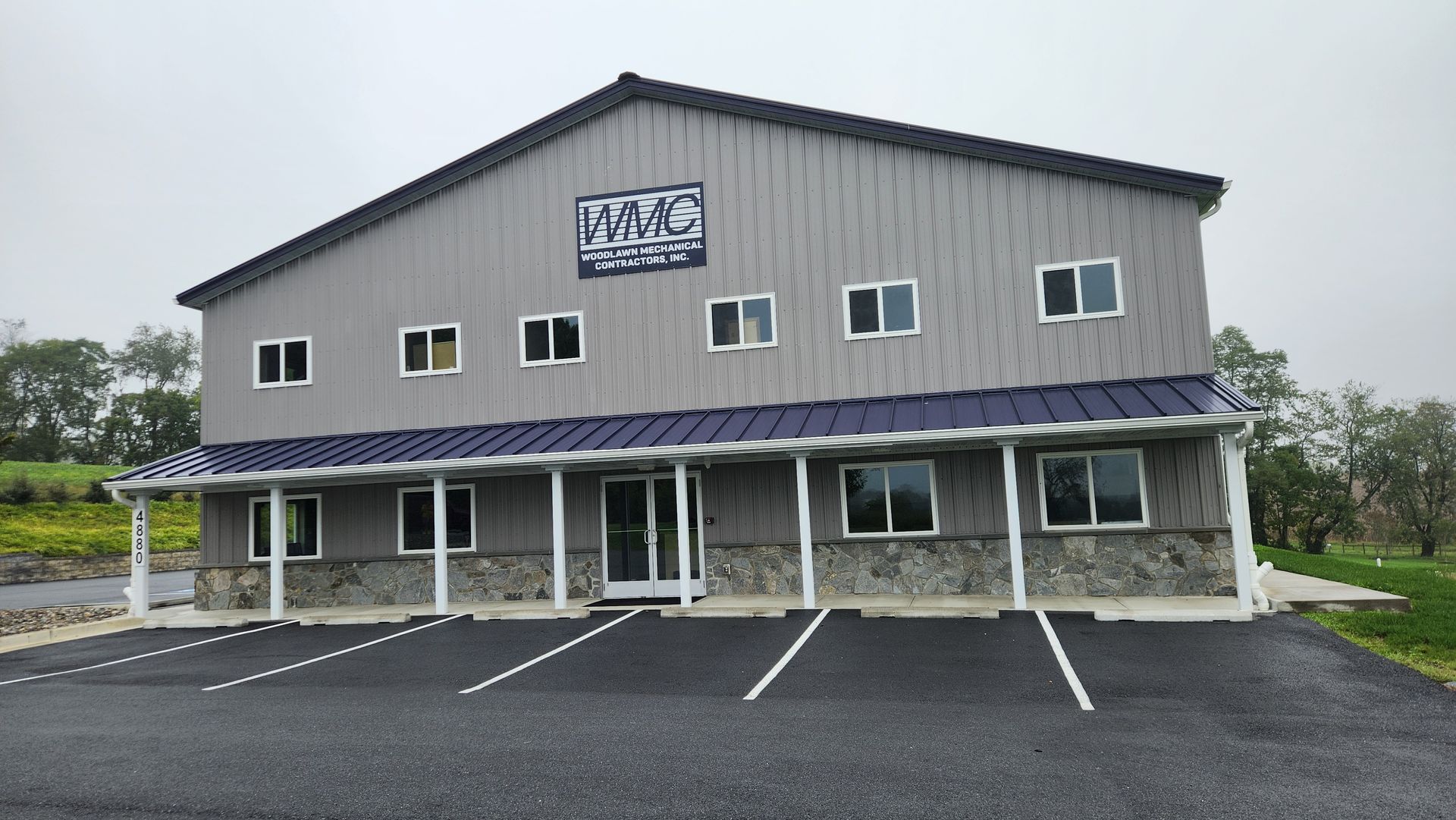 A two-story gray metal building with a stone base, blue roof trim, a centered entrance, and a paved parking lot.