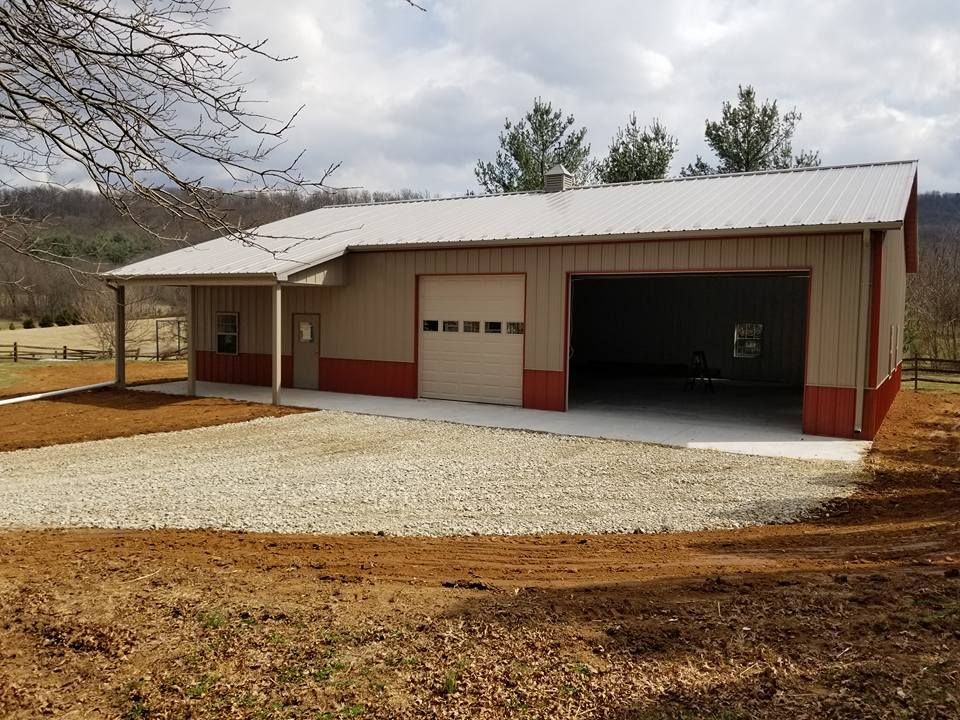 Tan and red metal garage with a covered porch and gravel driveway in a rural setting under a cloudy sky.