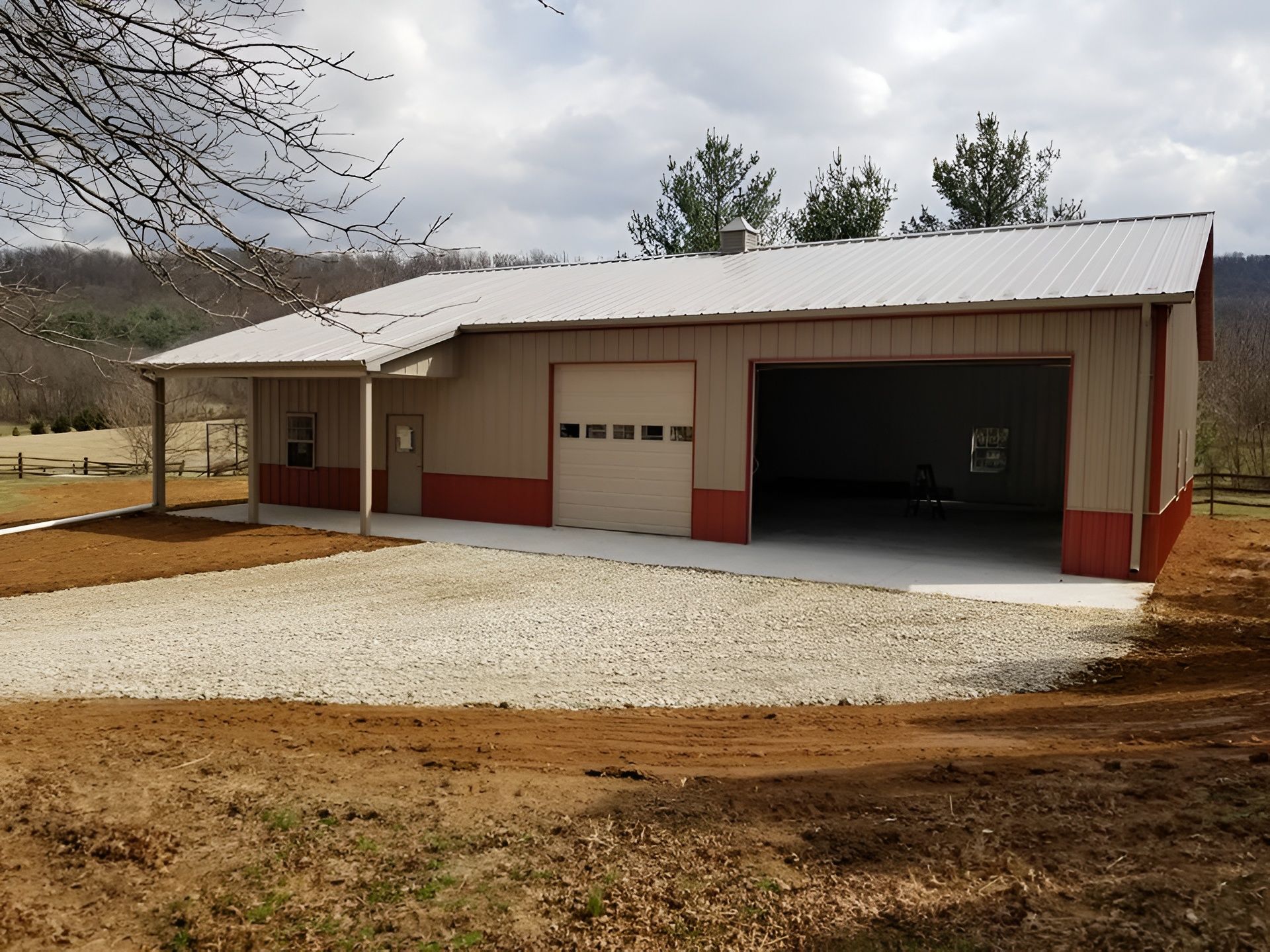 A tan metal pole barn with a red base, a front porch, a garage door, and an open bay, situated on a gravel lot.