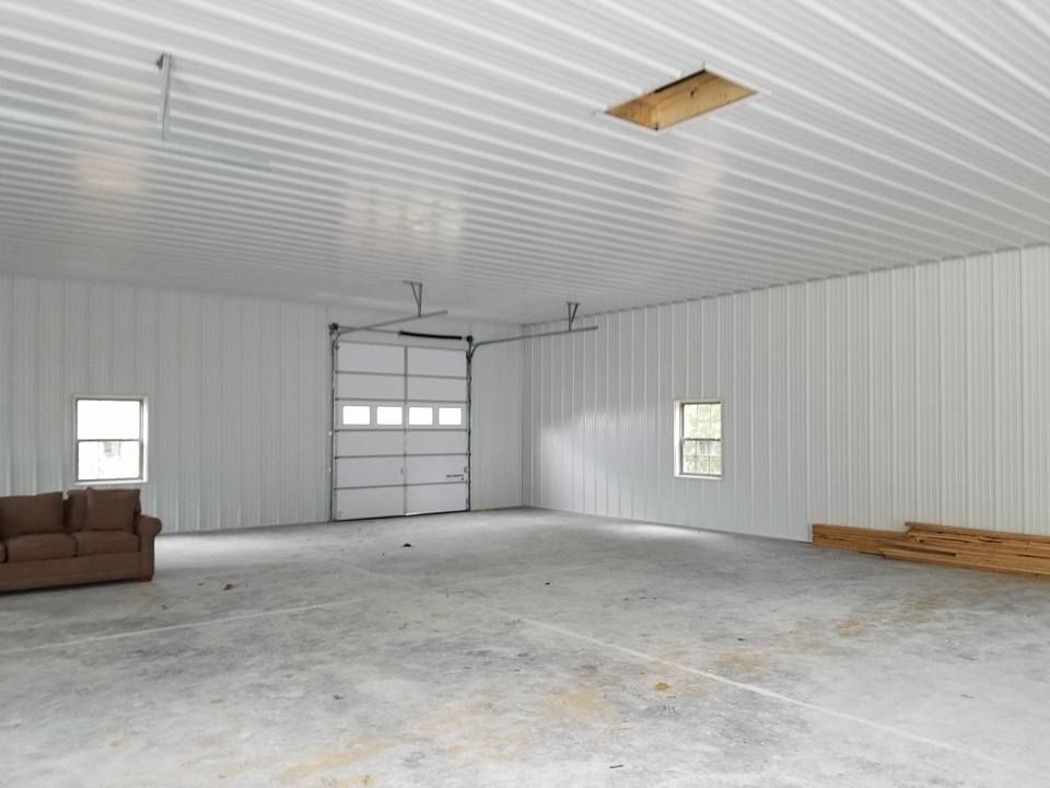 A spacious, white-walled metal garage with a concrete floor, a couch on the left, and a large central garage door.