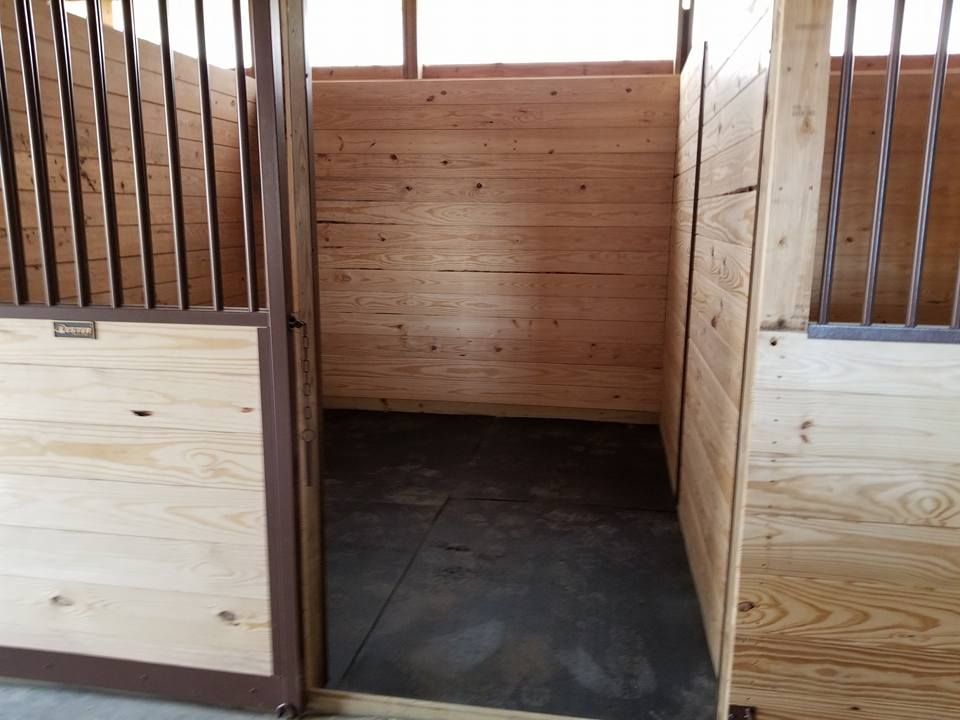 A view into an empty horse stall with light-colored wooden walls and a dark, solid floor.