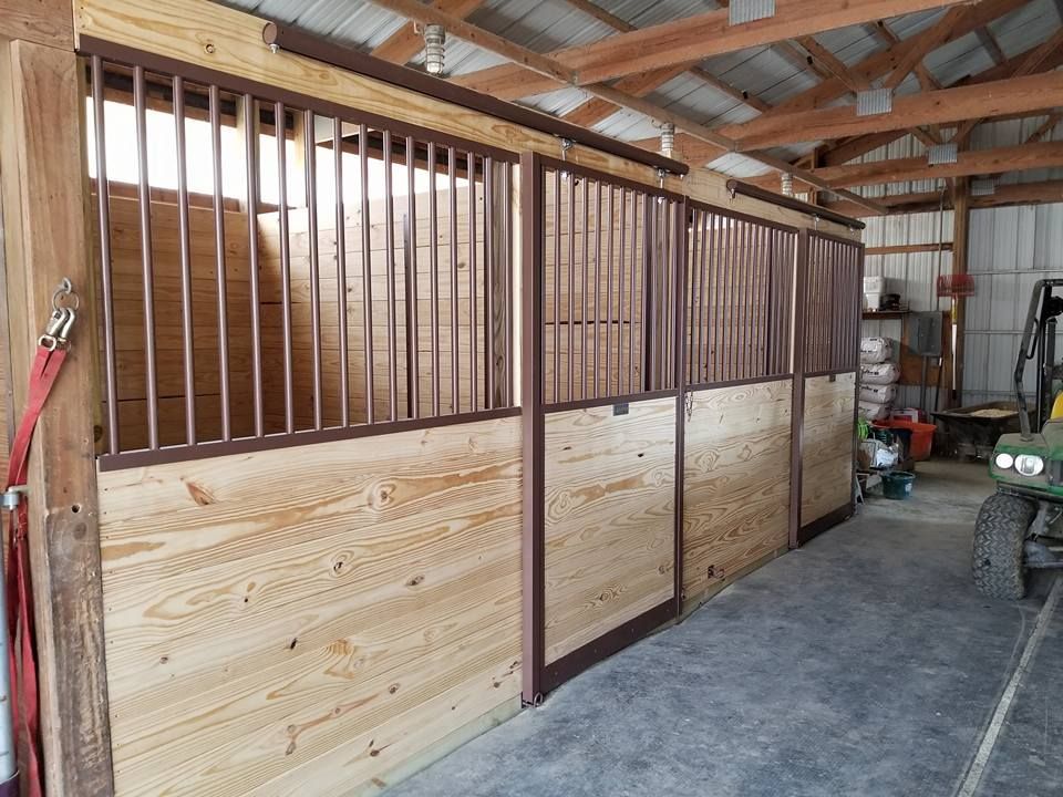 A row of horse stalls in a barn featuring wooden walls and black metal bars under an exposed timber ceiling.