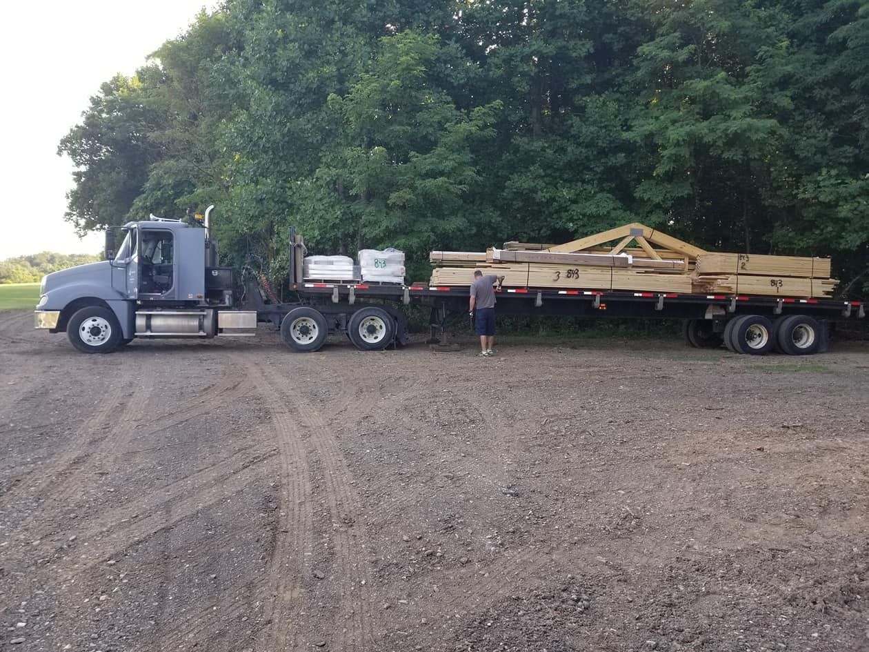A grey semi-truck with a flatbed trailer loaded with wooden beams and a truss parked on a gravel lot near trees.