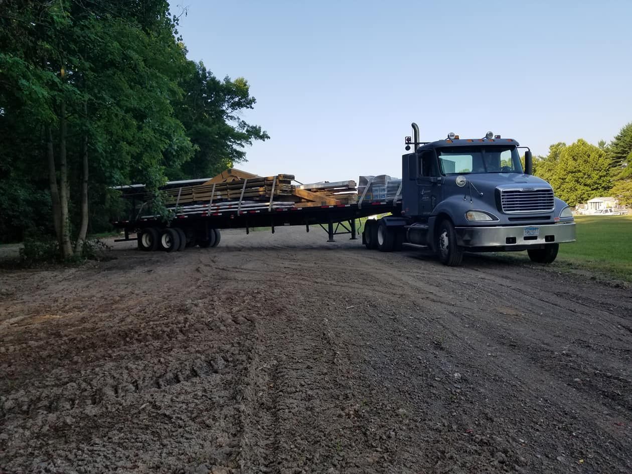 A gray semi-truck with a flatbed trailer loaded with lumber sits on a dirt lot near a line of trees.