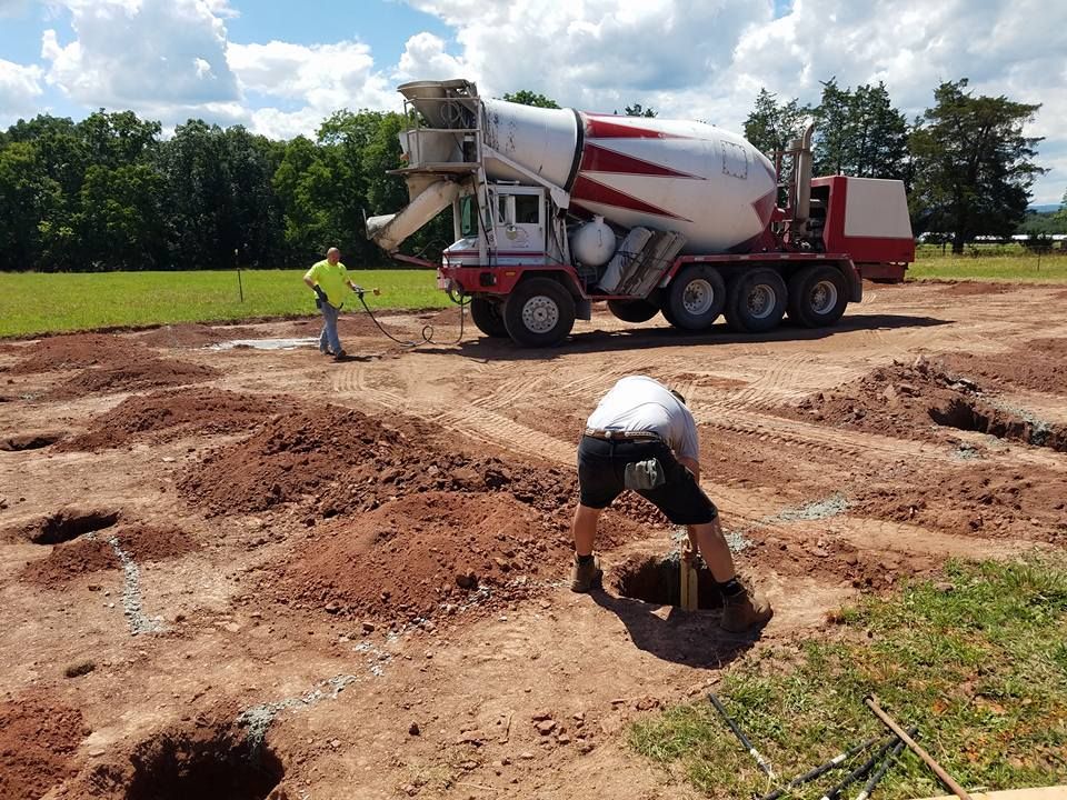 A worker stands by a concrete truck while another worker prepares a hole in a dirt construction site.