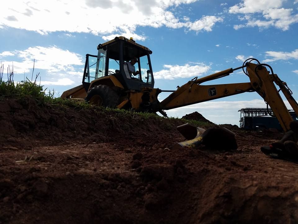 A yellow backhoe loader sits on a dirt pile under a bright, cloudy sky at a construction site.