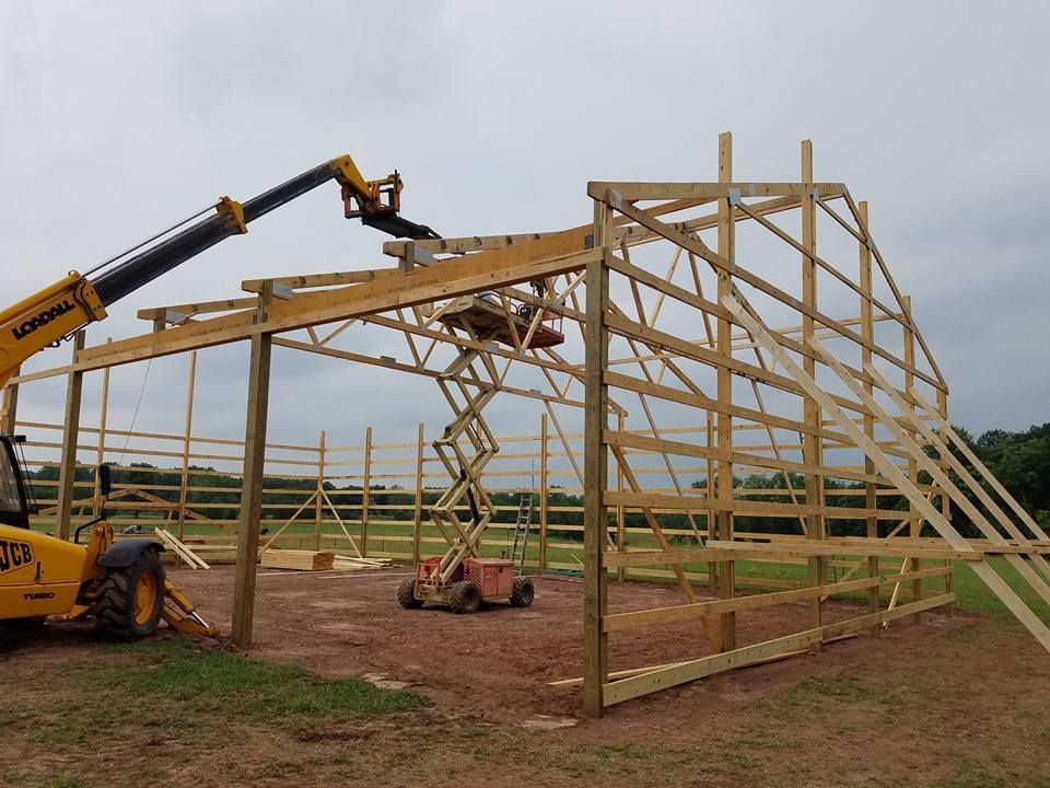 Construction workers use a telescopic handler and a scissor lift to build the wooden frame of a large pole barn.