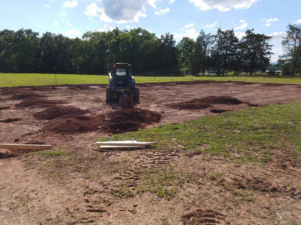 A tractor clears land on a sunny day, with piles of dirt scattered across a field bordered by trees.