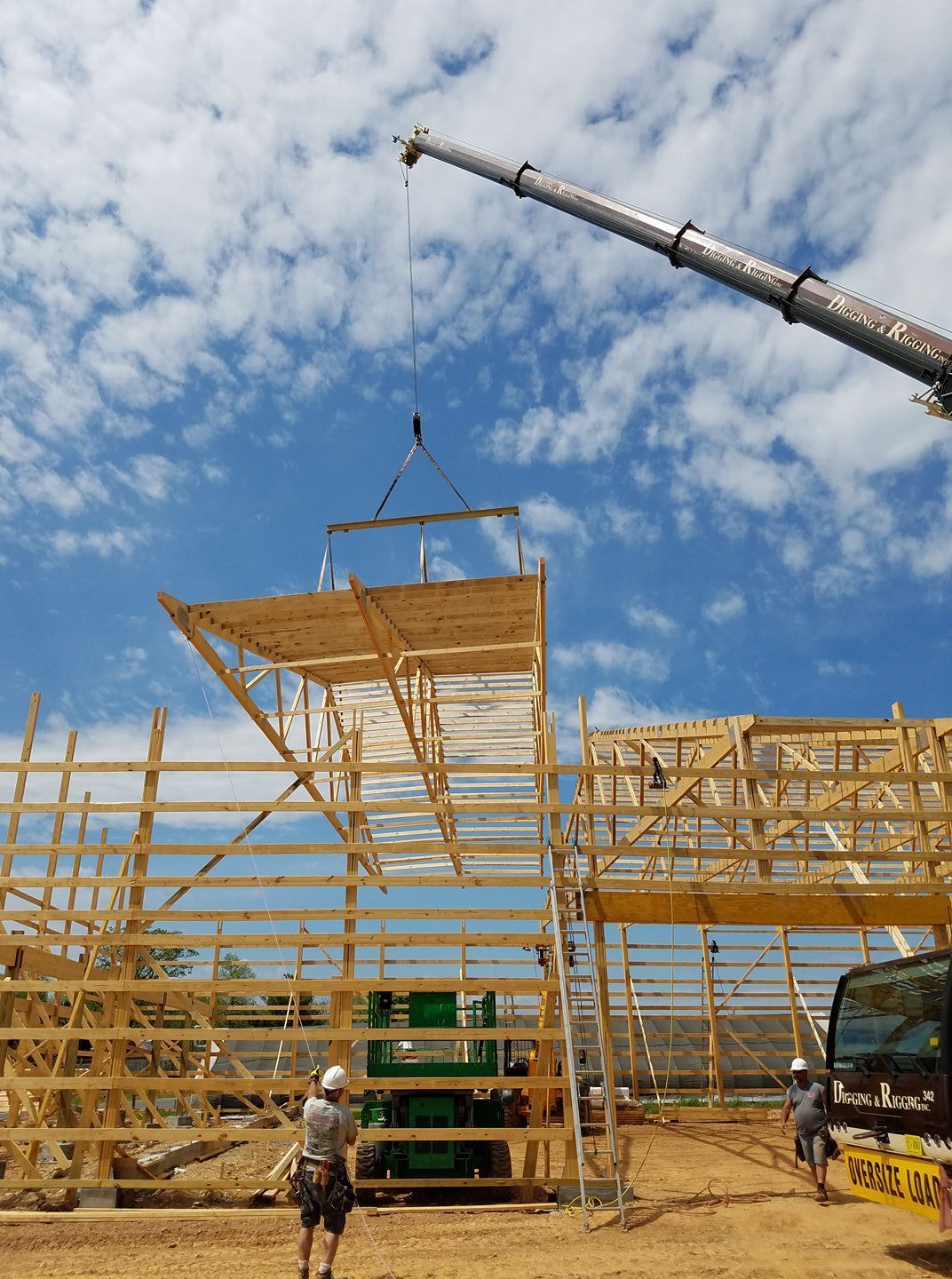 A crane hoists a large wooden truss structure into place at a construction site against a bright, cloudy blue sky.